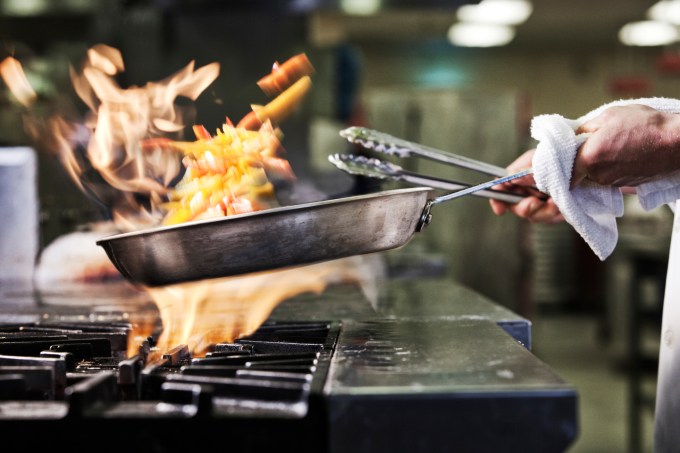 Nahaufnahme der Hände eines Kochs, der eine Bratpfanne hält, um Speisen zu kochen, wobei der Inhalt brennt. Flammen steigen aus der Pfanne.