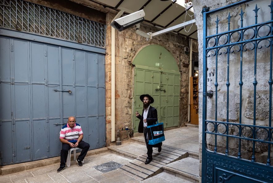 An ultra-Orthodox Jew passes through an empty alley in Jerusalem's Old City where a Palestinian is sitting in front of a closed store, November 2, 2023